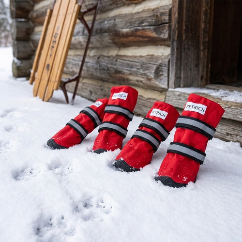 Botas rojas impermeables para perro sobre la nieve junto a una cabaña de madera